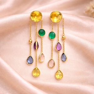 Può includere: Orecchini pendenti color oro con una grande pietra circolare gialla nella parte superiore. Ogni orecchino presenta una catena con una serie di pietre preziose colorate, tra cui forme a goccia verdi, viola, rosa, blu e gialle. Gli orecchini sono esposti su una superficie di raso rosa.