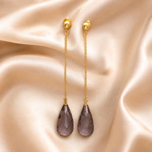 May include: A pair of gold-colored drop earrings with a teardrop-shaped, faceted, smoky quartz gemstone. The earrings hang from a small gold ball and chain. The background is a satin fabric.
