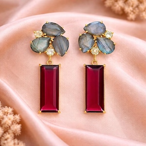 Può includere: Un paio di orecchini dorati con un gruppo di pietre grigio-blu a forma di goccia e piccole pietre trasparenti. Sotto, una grande pietra rettangolare rosso scuro pende da ogni orecchino. Presentati su raso rosa.