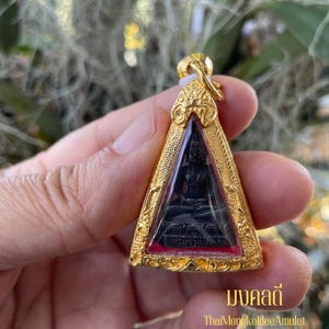 Puede incluir: Colgante dorado con un centro negro triangular que representa una figura sentada. El colgante tiene detalles ornamentados y un borde rojo. El texto "มงคลดี" y "ThaiMongkoldeeAmulet" son visibles en la parte inferior.