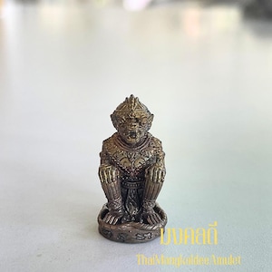 May include: A bronze-colored figurine of a seated, anthropomorphic figure with intricate detailing. The figure has a detailed headdress and ornate clothing. The base of the figurine is circular. The text "มงคลดี" and "ThaiMongkoldee Amulet" are visible.