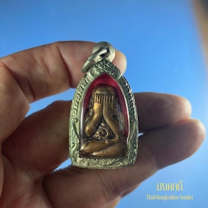 Puede incluir: Un amuleto de color bronce que representa una figura sentada con las manos cubriendo los ojos, enmarcada en un marco plateado ornamentado. El amuleto tiene un fondo rojo y un lazo para colgar. El texto "ThaiMongkoldeeAmulet" es visible.