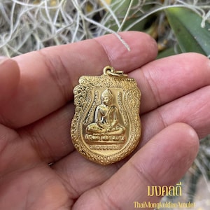 Puede incluir: Un colgante dorado con un diseño ornamentado que presenta una figura de Buda sentado. El colgante tiene forma de escudo con intrincados patrones y un lazo para colgar. El texto "มงคลดี" y "ThaiMongkoldeeAmulet" son visibles.
