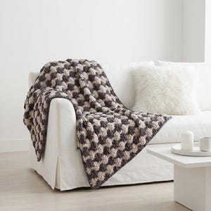 May include: A gray and beige patterned throw blanket draped over the arm of a white sofa. The blanket has a textured, raised design. A white fluffy pillow rests on the sofa. A small white table holds candles and cups.