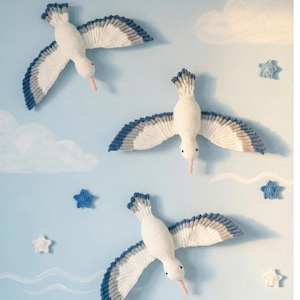 Può includere: Tre decorazioni murali a forma di gabbiano bianche all'uncinetto con accenti di ali blu e grigie sono esposte su uno sfondo azzurro con nuvole dipinte e decorazioni a forma di stella. I gabbiani hanno becchi rosa e occhi neri.