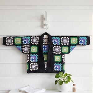 May include: A black crochet cardigan with a colorful patchwork design. The squares feature shades of green, purple, blue, and white. The cardigan is hanging on a white hanger against a white wall.