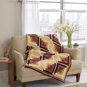 May include: A handmade crochet blanket draped over a beige armchair. The blanket features a geometric pattern with squares in shades of beige, yellow, orange, teal, and burgundy. The blanket is a cozy home decor item.
