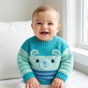 May include: A baby wearing a handmade crochet sweater. The sweater is blue and turquoise with a bear face design on the front. The bear face has a light blue nose and two dark blue eyes. The sweater has a round neck and long sleeves.