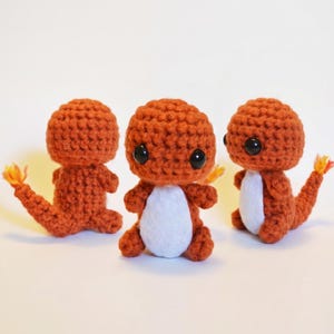 May include: Three handmade crochet Charmander figures. Each figure is orange with a white belly and black eyes. The tails have a yellow and orange flame. The figures are sitting on a white surface.