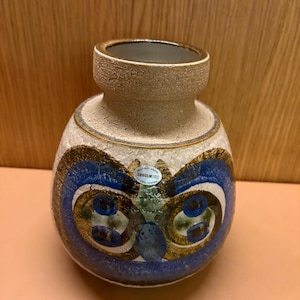 Puede incluir: Un jarrón de cerámica con un cuerpo beige moteado y texturizado y un borde marrón oscuro. El jarrón presenta un diseño abstracto azul, blanco y dorado. Una pequeña etiqueta con texto es visible en el lateral. El jarrón es un objeto decorativo.