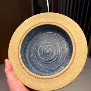 Op de afbeelding: Een handgemaakte keramische kom met een beige buitenkant en een blauwe binnenkant. De kom heeft een cirkelvorm met een getextureerd oppervlak en een bruine accentring. De binnenkant heeft een spiraalvormig ontwerp.