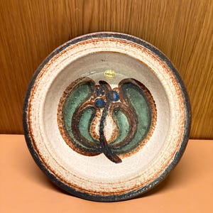 Könnte beinhalten: Eine Keramikschale mit cremefarbenem Innenraum und dunkelbraunem Rand. Die Schale hat ein abstraktes grünes und braunes Design in der Mitte. Das Äußere hat ein strukturiertes, erdiges Aussehen, was auf handwerkliche Qualität hindeutet. Ein kleiner Aufkleber ist sichtbar.