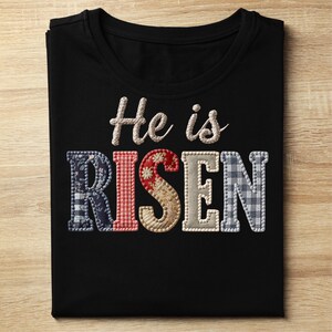 Può includere: T-shirt nera con la frase ricamata "He is RISEN". La parola "RISEN" è in stile patchwork con tessuto floreale, a quadri e fantasia. La maglietta è piegata su una superficie di legno chiaro.