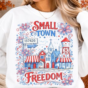 May include: A white sweatshirt featuring a graphic print with the text "SMALL TOWN FREEDOM" and "157426" on a sign. The design includes red, white, and blue illustrations of buildings, a water tower, and flowers, arranged in a patriotic colour scheme.