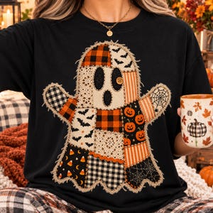 Może przedstawiać: Czarny t-shirt z dużą, centralną grafiką ducha wykonaną z różnych łatek materiału. Wzór ducha zawiera motywy pomarańczowo-czarnej kraty, nietoperzy, dyń, pajęczyn, koronki oraz cukierków w kształcie ziaren kukurydzy, z jednym guzikiem zamiast oka.