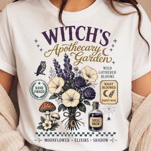 Op de afbeelding: Een wit t-shirt met een grafische print met de tekst "Witch's Apothecary Garden". Het ontwerp bevat een boeket paarse en witte bloemen, een uil, paddenstoelen, een spinnenweb en een fles met het label "Spell Brew" en botanische tekst.