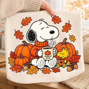 Può includere: Una borsa tote color crema con una grafica di Snoopy che indossa un maglione arancione e tiene una tazza con la scritta PUMPKIN SPICE. Il design include Woodstock, zucche arancioni, ghiande marroni e foglie autunnali su sfondo bianco.