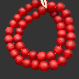 May include: A circular strand of opaque red beads with a matte, textured finish. The beads are strung together on a thin, light-colored cord, forming a continuous loop. Each bead has a slightly irregular, rounded shape with a consistent crimson hue.