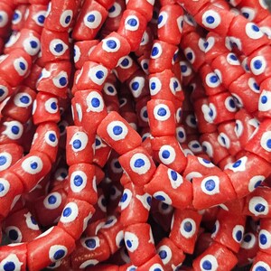 May include: A collection of cylindrical beads in a vibrant red color, each featuring a hand-painted eye motif. The design consists of a white circular center with a small, solid blue dot in the middle, repeated across every individual bead surface.