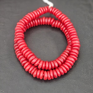 May include: A strand of small, circular, bright red beads strung together on a thin white cord. The beads are uniform in shape and size, featuring a smooth, matte finish, arranged in a continuous loop that forms a circular pattern.