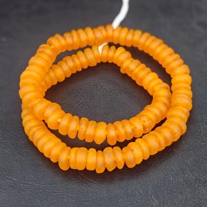 May include: A strand of small, round, translucent orange beads with a matte finish. The beads are strung on a thin white cord and arranged in a circular, double-layered loop, showcasing a consistent shape and vibrant, warm color throughout.