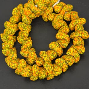 Könnte beinhalten: Ein kreisförmiger Strang aus kleinen, unregelmäßig geformten Perlen mit einer leuchtend gelben Grundfarbe und komplizierten Mustern aus dünnen, kurzen schwarzen Linien und orangefarbenen Strichen. Die Perlen sind auf einer weißen Schnur aufgereiht.