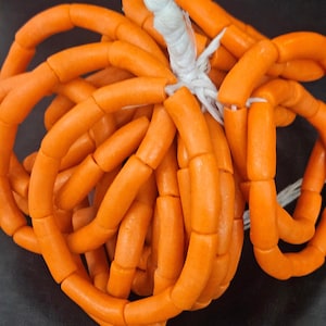 May include: A pumpkin-shaped decorative object constructed from numerous bright orange cylindrical beads strung together on white cord. The beads are arranged in vertical segments to mimic the rounded, ribbed appearance of a pumpkin.