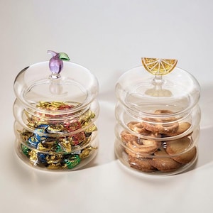 May include: Two clear glass jars with lids, one with a purple plum handle, the other a yellow lemon slice. The jars are filled with wrapped sweets and biscuits. The jars are stacked with three levels.