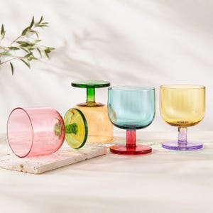 Puede incluir: Cuatro copas de vino de colores. Una es rosa, otra amarilla, otra azul y otra verde. Las copas están sobre una superficie blanca. Los tallos y las bases son de diferentes colores.