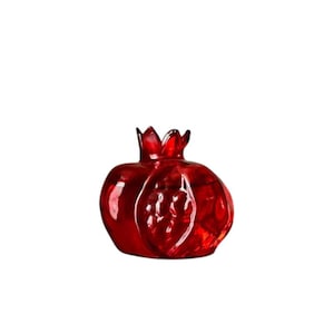 May include: A decorative, ruby-red glass pomegranate sculpture. The fruit-shaped ornament features a textured surface and a crown-like top. The translucent glass allows light to pass through, highlighting the rich color and details of the design.