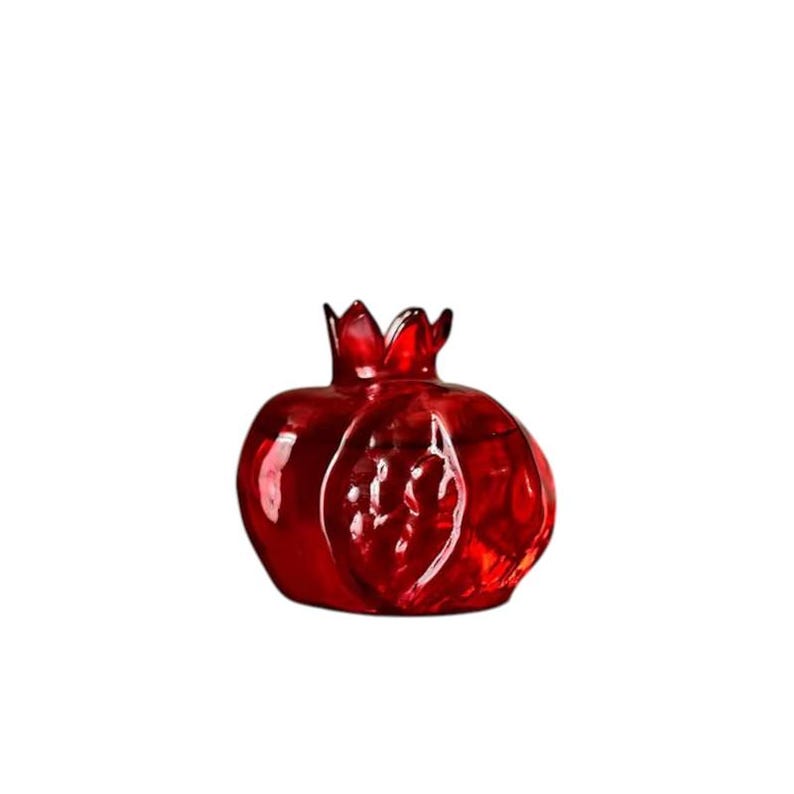 May include: A decorative, ruby-red glass pomegranate sculpture. The fruit-shaped ornament features a textured surface and a crown-like top. The translucent glass allows light to pass through, highlighting the rich color and details of the design.