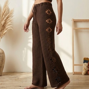 Puede incluir: Pantalones de crochet marrones con cintura con cordón y recortes florales. Los pantalones de pernera ancha están hechos de un material tejido texturizado. Los recortes florales están a lo largo del lateral. Los pantalones son de color marrón liso.