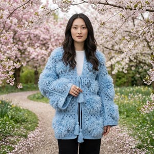 May include: A light blue crochet cardigan featuring a textured granny square pattern with integrated fuzzy, shaggy yarn accents. The garment has long, wide sleeves, a straight hem, and a front tie closure finished with two long, dangling tassels.