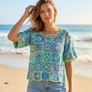 May include: A colorful crochet top with a square pattern in shades of blue, green, and purple. The top has short sleeves and a square neckline. The model is wearing blue jeans and is on a beach.
