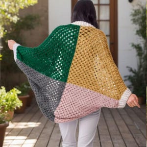 Op de afbeelding: Een kleurrijke, open geweven gehaakte poncho in groen, goud, grijs en roze. De poncho heeft een vierkant patroon en witte afwerking rond de kraag en manchetten. De persoon draagt een witte broek.