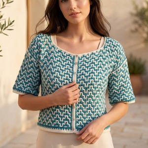 Puede incluir: Cárdigan con estampado en color turquesa y blanco, con mangas cortas y cierre de botones. El suéter presenta un diseño geométrico y un ribete color crema alrededor del cuello, las mangas y el dobladillo. El cárdigan es de punto suave.