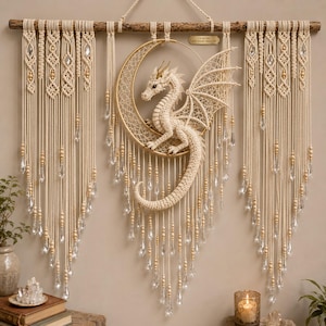 May include: A macrame wall hanging featuring a central cream-colored dragon perched on a gold crescent moon. The piece is suspended from a wooden branch and decorated with intricate knot patterns, wooden beads, and clear crystal-like pendants.