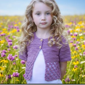 May include: A lavender crochet cardigan with short sleeves and wooden buttons. The bottom of the cardigan features a decorative openwork design. The cardigan is worn over a white top. The background is a field of colorful wildflowers.