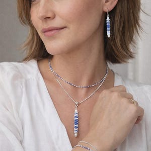 Op de afbeelding: Een vrouw draagt een gelaagde ketting, oorbellen en een armband. De sieraden hebben een patroon van blauwe en witte kralen. De ketting heeft een verticale hanger. De oorbellen zijn bungelend. De armband is een dubbele streng. De vrouw draagt een witte blouse.