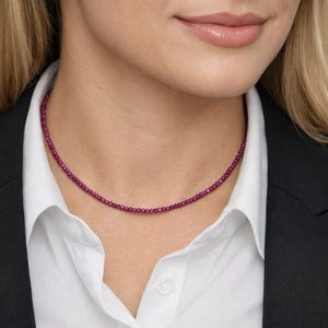 Könnte beinhalten: Eine zarte Rubin-Halskette, bestehend aus kleinen, runden, tiefroten Edelsteinen, wird zu einem weißen Hemd mit Kragen und einem schwarzen Blazer getragen. Die Halskette liegt eng am Hals an. Nahaufnahme.