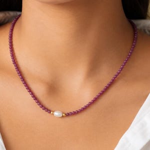 Puede incluir: Un collar que presenta una hilera de pequeñas cuentas de color magenta intenso. El collar está centrado con una sola perla blanca de forma ovalada y lisa, flanqueada por dos diminutas cuentas de metal en tono dorado.