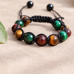 May include: A beaded bracelet featuring alternating round polished stones in shades of deep red, golden brown, and emerald green. The stones are connected by a black woven cord with an adjustable sliding knot closure and two small black beads.