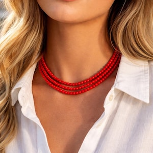 Puede incluir: Un collar de cuentas rojo de tres hilos. El collar está compuesto por pequeñas cuentas redondas y se lleva sobre una camisa blanca abotonada. La imagen es un primer plano del collar y del cuello y el pecho de la persona.