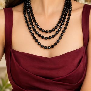 May include: A multi-strand necklace featuring three layers of smooth, round, glossy black beads. The necklace is worn against a dark burgundy sleeveless dress with a draped, cowl-style neckline that sits across the chest.