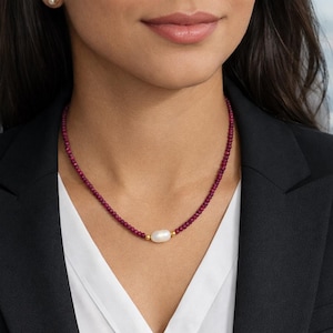 Könnte beinhalten: Eine Halskette mit kleinen, tiefroten Perlen und einer zentralen, ovalen Perle. Die Perle ist mit goldfarbenem Metall akzentuiert. Die Halskette wird über einem weißen Hemd und einem schwarzen Blazer getragen.
