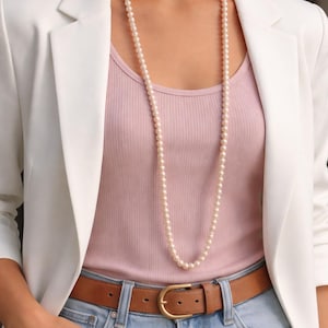 May include: A long strand of cream-colored pearls worn over a pink ribbed tank top. The necklace is paired with a white blazer, blue jeans, and a brown leather belt with a gold buckle. The outfit is casual and stylish.