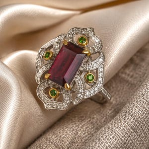 Puede incluir: Un anillo de tono plateado con una gran piedra preciosa rectangular de color rojo intenso engastada en garras doradas. El anillo está rodeado por un marco calado intrincado incrustado con pequeñas piedras claras y cuatro pequeñas piedras verdes redondas.