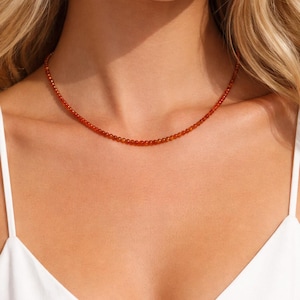 Puede incluir: Un delicado collar compuesto por pequeñas cuentas redondas de color rojo anaranjado. El collar se lleva alrededor del cuello, descansando sobre el pecho. Las cuentas están espaciadas uniformemente, creando un diseño simple pero elegante. El fondo es una blusa blanca.