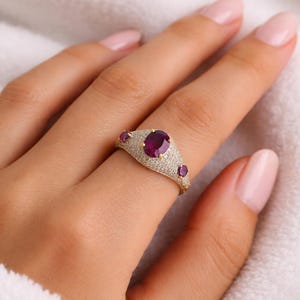 May include: A gold ring featuring a large, oval-cut ruby center stone, surrounded by smaller diamonds. Two smaller rubies flank the center stone. The ring is set on a white background.