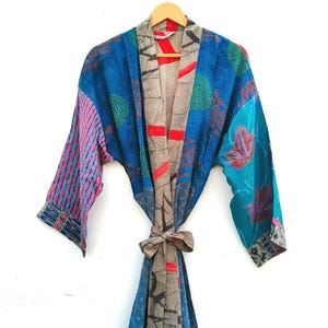May include: A long-sleeved kimono-style robe featuring a patchwork design with various blue, pink, and tan fabric panels. The garment includes a tan-coloured front placket and a matching tie belt, decorated with abstract patterns and floral motifs.